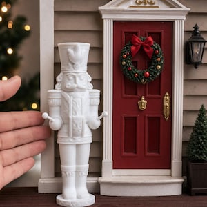 May include: A white 3D-printed nutcracker figurine standing in front of a miniature house facade with a red door, a small green wreath with red and gold ornaments, a gold door knocker, a gold handle, and a black wall-mounted lantern light fixture.