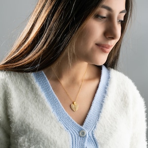 May include: A woman wearing a white and blue cardigan with a gold heart-shaped pendant necklace.