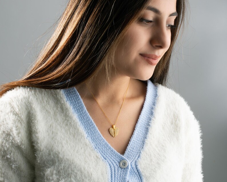 May include: A woman wearing a white and blue cardigan with a gold heart-shaped pendant necklace.