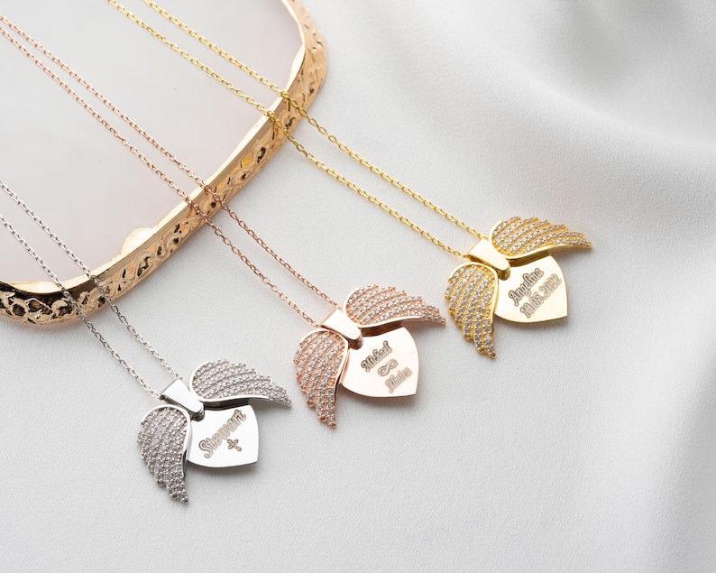 May include: Three silver necklaces with angel wing pendants. Each pendant features a heart-shaped charm with a name and date engraved on it. The names are "Michael", "Sophia", and "Angelina Marie 2021".