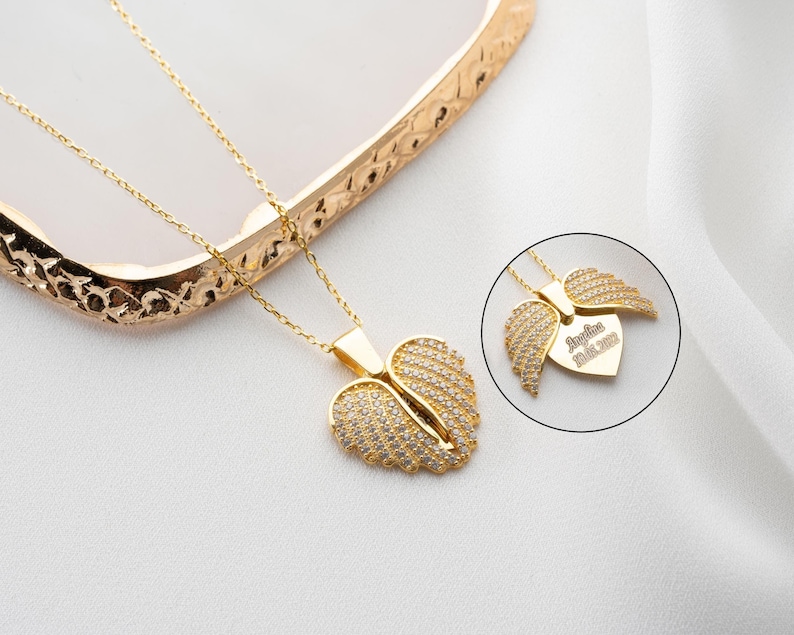 May include: Gold necklace with a heart-shaped pendant featuring two angel wings with clear cubic zirconia stones. The pendant has an inscription that reads "Angelina 01.01.2022".