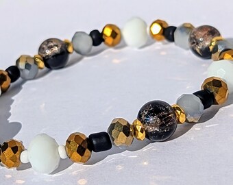 May include: A bracelet made of black, white, gold, and gray beads. The beads are of various shapes and sizes, including round, square, and rectangular.