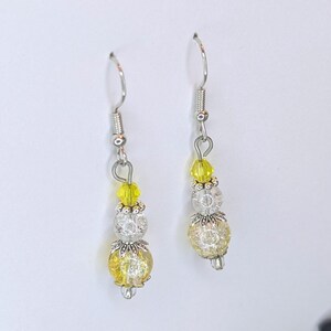 May include: A pair of dangle earrings with silver-tone hooks. Each earring features a yellow, clear, and silver bead design. The earrings have a floral-like design on the silver beads and a yellow crystal bead at the top.