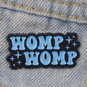 Può includere: Uno spillo in smalto nero con il testo "WOMP WOMP" in lettere blu con stelle bianche su uno sfondo in denim.