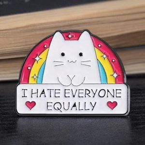 May include: A white cat-shaped enamel pin with a rainbow background and the text "I HATE EVERYONE EQUALLY." The pin features a white cat face, a rainbow, and two red hearts. The pin is a fun accessory.
