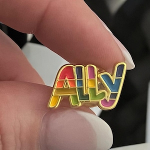 以下が含まれることがあります： 虹色の「ALLY」という文字が入った金色のピンバッジ。2本の指でピンバッジを持っています。文字は赤、オレンジ、黄色、緑、青、紫で塗りつぶされています。