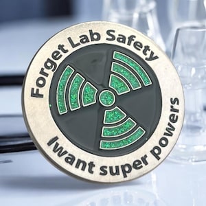 May include: A circular metal coin with the words "Forget Lab Safety" and "I want super powers" around the edge. The centre has a green, glittery radiation symbol on a dark grey background.