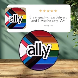 以下が含まれることがあります： 虹色のデザインと黒い文字で「ally」と書かれた丸いピン。ピンは木製の表面にあります。