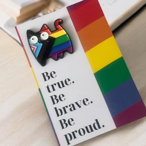 Puede incluir: Un marcapáginas con una franja de colores del arcoíris y un pin de gato con un diseño de arcoíris y un cuerpo negro. El marcapáginas tiene las palabras "Be true. Be brave. Be proud." impresas.