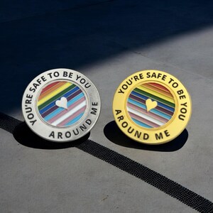 Könnte beinhalten: Zwei runde Anstecknadeln mit dem Text "YOU'RE SAFE TO BE YOU AROUND ME". Eine Nadel ist silberfarben mit einem Regenbogen-Design und einem weißen Herzen. Die andere ist goldfarben mit einem Regenbogen-Design und einem goldenen Herzen.