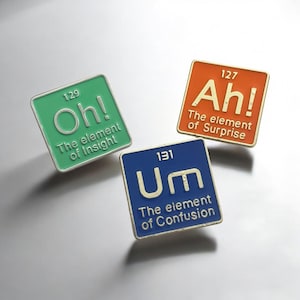 May include: Three square enamel pins with a metallic border. The pins are green, orange, and blue, each with a different expression: "Oh!", "Ah!", and "Um". Each pin has text that reads "The element of..." followed by a word.