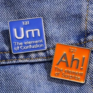 May include: Two novelty pins shaped like periodic table elements. The blue pin says "Um" with the atomic number 131 and "The element of Confusion". The orange pin says "Ah!" with the atomic number 127 and "The element of Surprise".