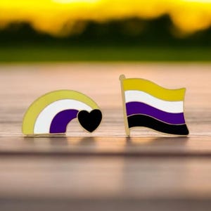 May include: Two enamel pins. One is a rainbow in yellow, white, and purple with a black heart. The other is a flag with yellow, white, purple, and black stripes. Both pins have gold-coloured borders.