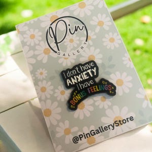 May include: Two enamel pins on a white card with a daisy pattern. The top pin is black with white text that reads "I don't have anxiety I have bonus feelings". The bottom pin is rainbow with black text that reads "bonus feelings".