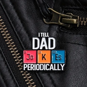 Op de afbeelding: Een zwart leren jack met een rits en een pin die zegt "I tell dad jokes periodically". De pin heeft drie kleurrijke vierkanten met de letters "Jo", "K" en "Es" erop, die de chemische elementen voorstellen.
