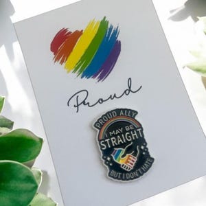 Puede incluir: Una tarjeta blanca con un corazón arcoíris y la palabra "Proud" escrita en cursiva negra. Un pin de esmalte negro con un arcoíris y el texto "Proud Ally I May Be Straight But I Don't Hate" está en la tarjeta.