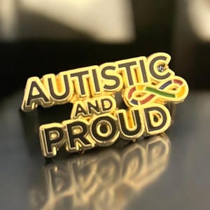Könnte beinhalten: Ein goldfarbener Ansteckpin mit den Worten "AUTISTIC AND PROUD" in schwarzer Schrift. Der Pin zeigt ein farbenfrohes Unendlichkeitszeichen, das Neurodiversität repräsentiert. Der Pin hat eine glänzende, reflektierende Oberfläche.