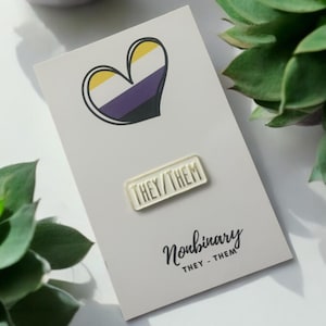 May include: A white card featuring a non-binary heart symbol and a pin reading "THEY/THEM." The card also has the word "Nonbinary" written on it. Green succulents are in the background.