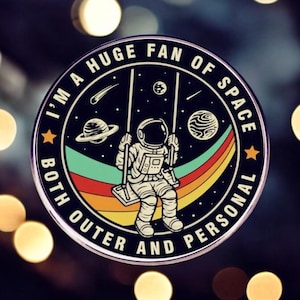 May include: A round, silver pin with a black background and colourful rainbow design. The pin features an astronaut on a swing in space with the text "I'm a huge fan of space, both outer and personal."