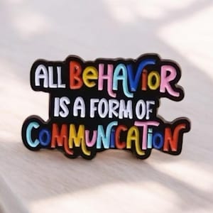 Puede incluir: Pin de esmalte negro con la frase "ALL BEHAVIOR IS A FORM OF COMMUNICATION" en letras coloridas estilo burbuja. El pin tiene un borde negro y está sobre una superficie clara.