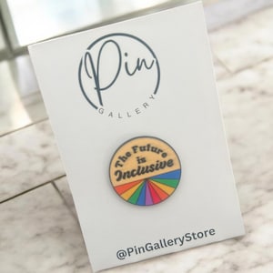 May include: A round wooden pin with a rainbow design and the text "The Future is Inclusive". The pin is on a white card with the text "Pin Gallery" and "@PinGalleryStore".