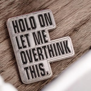 May include: A silver-coloured pin featuring the text "HOLD ON LET ME OVERTHINK THIS" in black, block lettering. The pin has a rectangular shape with a distinctive cutout design. The pin is set against a wooden background.