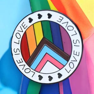 May include: A circular enamel badge with a white outline, black hearts, and the words "LOVE IS LOVE." The badge features a rainbow peace symbol with LGBTQ+ pride flag colours. The background is a rainbow flag.