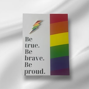 以下が含まれることがあります： 「Be true. Be brave. Be proud.」というテキストと虹色の稲妻のピンが付いた白いカード。カードは虹色のストライプの背景になっています。