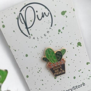 May include: A green cactus enamel pin with a black pot that says "Don't be a prick". The pin is on a white background with green splatter paint.
