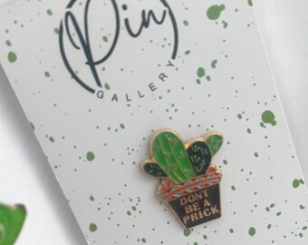 Puede incluir: Un pin de esmalte de cactus verde con una maceta negra que dice "Don't be a prick". El pin está sobre un fondo blanco con salpicaduras de pintura verde.