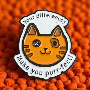 May include: A cat-shaped enamel pin with an orange body, brown ears, and a white border. The pin features the text "Your differences Make you purr-fect!" in black lettering. The cat has a unique eye design.