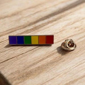 May include: A rectangular enamel pin with a rainbow design, representing the LGBTQ+ pride flag. The pin has a gold-coloured backing and a pin clasp. The pin is displayed on a wooden surface.