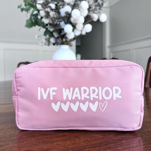 May include: A pink zippered pouch with the text "IVF Warrior" and a row of white hearts printed on the front.