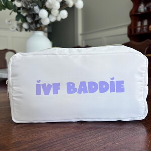 May include: White zippered pouch with the text "IVF BADDIE" in purple lettering with small hearts on either side of the text.
