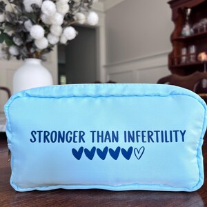 May include: A light blue rectangular cosmetic bag with a zipper closure. The bag features the text "STRONGER THAN INFERTILITY" in dark blue, along with several heart symbols. The bag is sitting on a wooden surface.