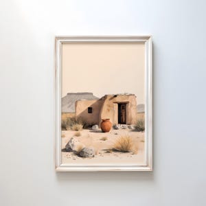 May include: Framed print of a desert landscape featuring a weathered adobe structure with a wooden door and a terracotta pot. The artwork depicts a serene scene with a mountain in the background, set against a pale beige sky.