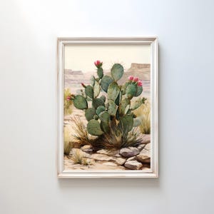 Op de afbeelding: Een ingelijste aquarel van een cactus met roze bloemen in een woestijnlandschap. De groene cactus staat centraal, omringd door droog gras en rotsen. De achtergrond toont mesas en een lichte lucht, alles omlijst door een wit houten frame.