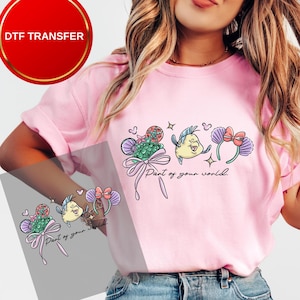 May include: Pink t-shirt featuring a colorful graphic of cartoon characters and the text "Part of your world." The design includes a fish, a seashell, and a bow. The shirt is a DTF transfer.