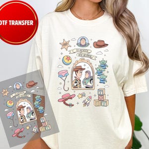 May include: Cream-colored t-shirt featuring a colorful graphic design with characters and elements from the animated film "Toy Story." The design includes Woody, Buzz Lightyear, aliens, and the phrase "To Infinity and Beyond."