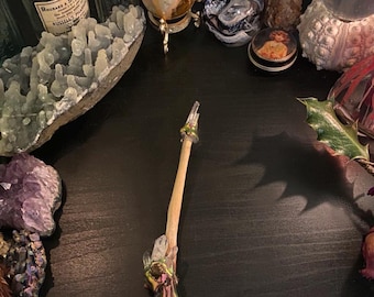 May include: A wooden wand with clear quartz crystals and green accents. The wand is lying on a dark surface.