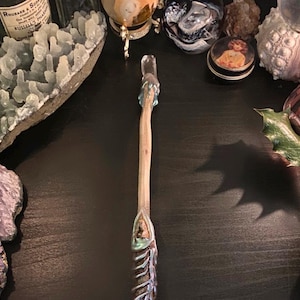 Pode incluir: Uma varinha de madeira com um cristal iridescente azul claro e verde no topo. A varinha está sobre uma superfície escura com outros objetos ao redor.