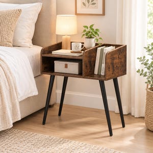 May include: A dark brown wood-grain nightstand featuring four black tapered legs, an open storage shelf, and a vertical divider. The top surface holds a small lamp, a white mug, and a potted plant, while the shelf contains a storage box and books.