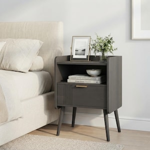 May include: A dark gray wooden nightstand with four tapered legs, an open storage shelf, and a single drawer featuring a gold-toned handle and vertical grooved detailing. The top surface holds a framed photograph, a small bowl, and a green plant.