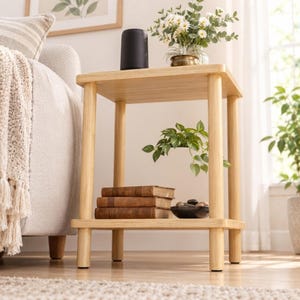 May include: A light-toned wooden side table with a square top and a lower shelf. The top surface holds a black cylindrical object and a small brass pot with white flowers. The lower shelf contains three stacked brown books and a dark bowl.