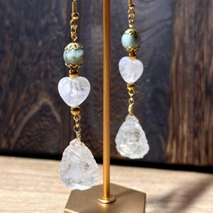 May include: Gold-toned dangle earrings with a natural, earthy aesthetic. Each earring features a gold hook, a green-grey bead, a heart-shaped clear crystal, and a rough-cut clear crystal pendant. The earrings are displayed on a gold stand.