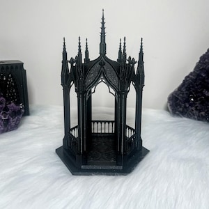 Puede incluir: Un gazebo de estilo gótico negro con detalles intrincados y un techo puntiagudo. El gazebo está hecho de un material oscuro y texturizado y tiene un pequeño piso abierto.