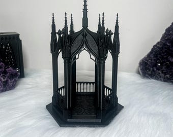 May include: A black gothic-style gazebo with intricate details and a pointed roof. The gazebo is made of a dark, textured material and has a small, open floor.