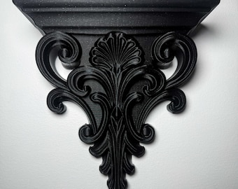 May include: Black wall shelf with ornate, scrollwork detailing. The shelf has a flat top and a decorative, symmetrical design. The shelf is mounted on a white wall, creating a contrast with the dark color of the shelf.