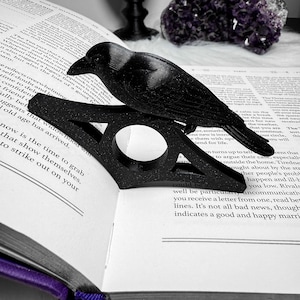 May include: A black crow ornament sits on a black book page holder, placed on an open book. The book is open to a page of text. The crow and holder are made of a matte material. The scene is set on a white surface.