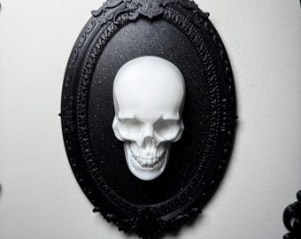 Op de afbeelding: Een witte schedel in een zwart, sierlijk ovaal frame. Het frame is bevestigd aan een witte muur. De schedel is het centrale punt en suggereert een gotische of Halloween-esthetiek. De details van het frame voegen een decoratief element toe.